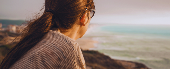 Woman with ponytail looking out over the ocean from a cliff, appearing thoughtful and reflective.