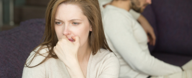 Woman sitting in distress while partner sits turned away in the background, illustrating emotional distance and relationship tension
