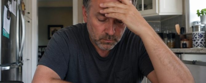 Man sitting at kitchen table holding his head, showing stress and anxiety that can lead to addiction